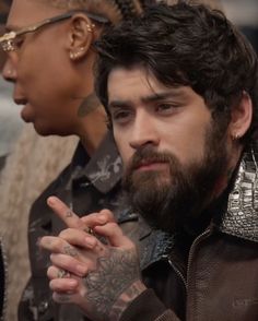 a bearded man with tattoos and piercings sitting next to another person in front of him