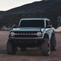 the front end of a gray truck parked on top of a dirt covered field with mountains in the background