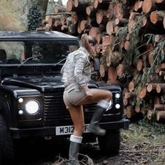 a woman sitting on the hood of a black land rover parked in front of a pile of logs