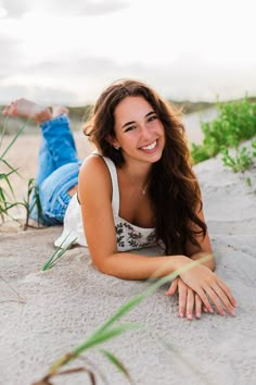 a beautiful young woman laying on top of a sandy beach next to tall grass and plants