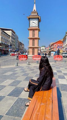 a woman sitting on top of a wooden bench next to a clock tower in the background