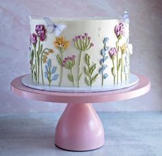 a white cake with colorful flowers and butterflies on it sitting on a pink stand in front of a wall