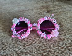 a pair of pink sunglasses with flowers and pearls on the rim, sitting on a wooden surface