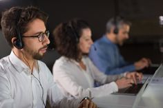 three people sitting at a desk with laptops and headphones