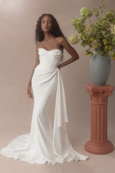 a woman in a white dress standing next to a vase with flowers and greenery