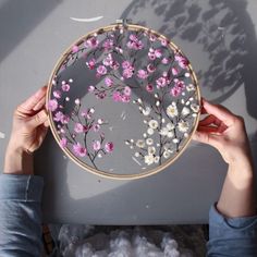 a person holding up a embroidery project with pink and white flowers on it, in front of a gray background