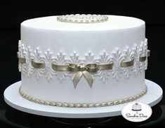 a white wedding cake with gold ribbon and bow on the top is sitting on a glass platter