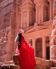 a woman in a red dress sitting on the side of a cliff near an ancient building