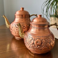 two metal teapots sitting on top of a wooden table next to a potted plant