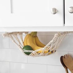 bananas and other fruits are in a white basket on the kitchen counter next to a wooden spoon