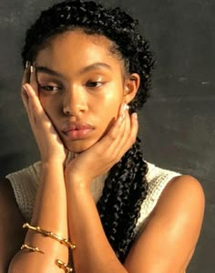 a woman with her hands on her face and wearing gold bracelets, standing in front of a black background