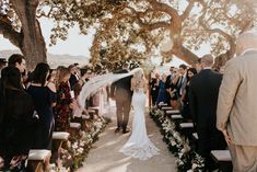 a bride and groom walking down the aisle at their outdoor wedding ceremony under a large tree