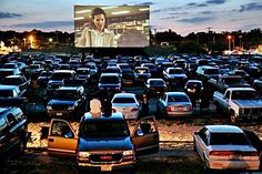 a large group of cars parked in a parking lot with a movie screen behind them