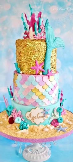 a three tiered cake decorated with gold, blue and pink mermaid tail decorations on top