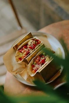 there are three tacos on the plate and one is wrapped in brown paper with toppings