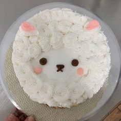 a cake with white frosting and pink ears is on a clear platter while someone holds their hand out to it