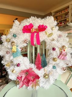 a white wreath with pink bows and ornaments on top of a table in front of a mirror