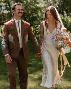 a bride and groom holding hands walking through the grass with flowers in their bouquets