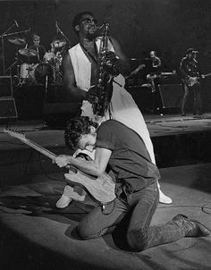 a man kneeling down next to another man with a guitar in his hand and other people on the stage behind him