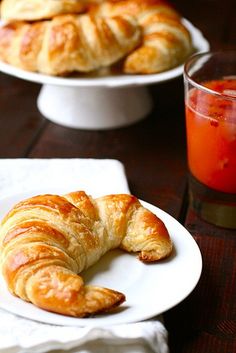 croissants on white plates next to a glass of juice and bowl of fruit