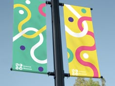 two colorful banners hanging from a pole on top of a street light post with trees in the background