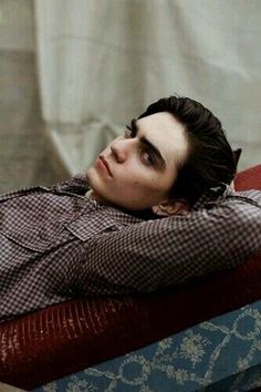 a young man laying on top of a bed next to a red chair and white curtain