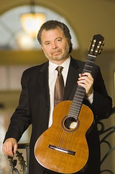 a man wearing a suit and tie holding a guitar in his right hand while standing next to a railing