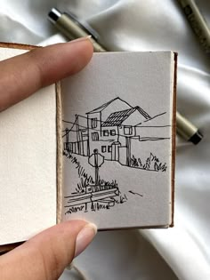 a hand is holding a small piece of paper with a house drawn on it and two pens are in the background