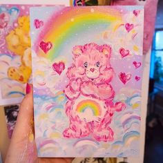 a hand holding up a card with a pink teddy bear and rainbow in the background