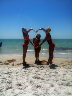 three women in bikinis standing on the beach making a heart shape with their hands