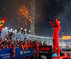 a man in a red race suit standing on top of a car with fireworks behind him