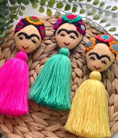 three little dolls with tassels sitting on a woven basket next to some plants