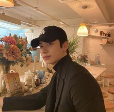 a young man sitting at a table with flowers in a vase on the counter behind him