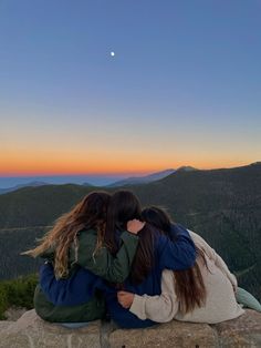 two women sitting on top of a rock with their arms wrapped around each other looking at the sunset