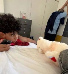 a young boy laying in bed next to a stuffed animal and holding a remote control