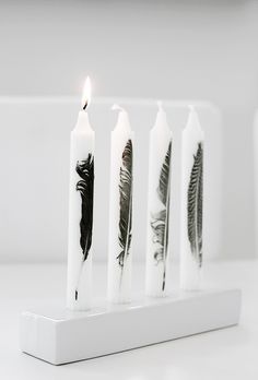 four candles with black feathers on them sitting in front of a white wall and one candle is lit