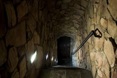 a long narrow stone tunnel with light coming through the door and on the wall there is a metal handrail