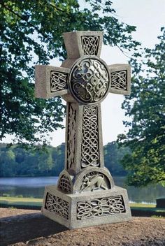 a stone cross sitting in the middle of a field next to trees and water on a sunny day