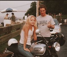 a man standing next to a woman sitting on a motorcycle