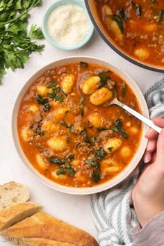 a person holding a spoon in a bowl of stew with bread and parsley on the side