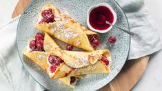 three crepes with berries and powdered sugar on a plate next to a cup of syrup