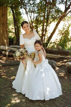 two women in wedding dresses standing next to each other on the grass with trees behind them