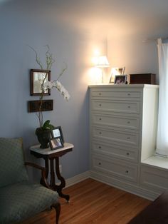 a room with a chair, dresser and lamp on the side table in front of a window