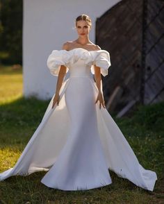 a woman in a white wedding dress standing on the grass with her arms behind her back