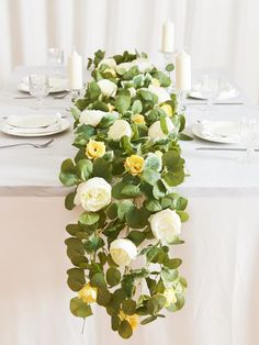 the table is set with white and yellow flowers, greenery and candles on it