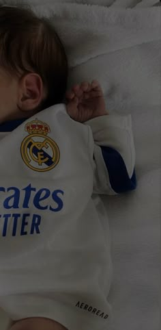 a baby laying on top of a bed wearing a white shirt with the real madrid logo