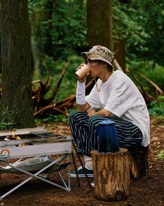 a man sitting on a stump drinking from a cup next to a campfire in the woods