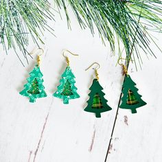 three green christmas trees are hanging from gold earwires on a white wooden table