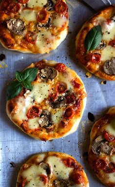 four small pizzas with cheese, mushrooms and basil on them sitting on a piece of parchment paper