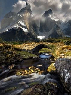 a stream running through a lush green field next to rocks and grass with mountains in the background